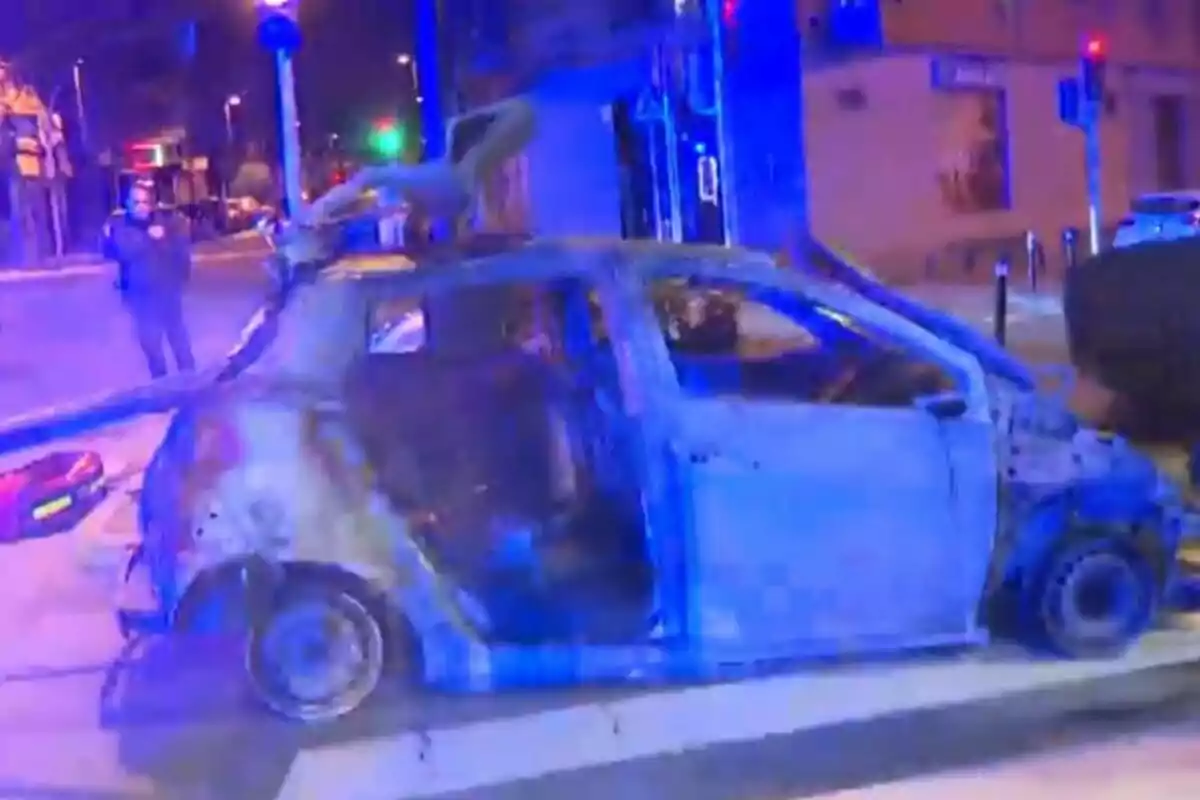 Un coche quemado en una intersección iluminada por luces azules durante la noche.