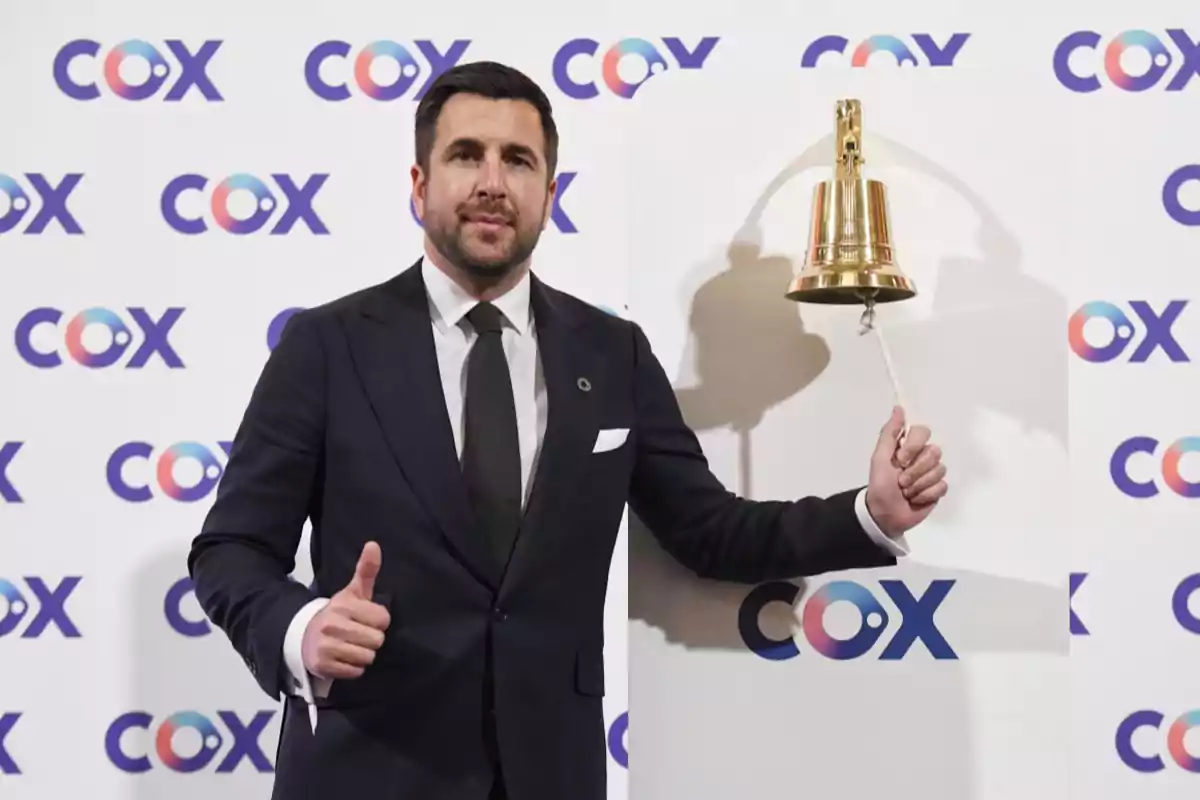 Man in a dark suit smiling and giving a thumbs up while holding a golden bell in front of a background with the COX logo.