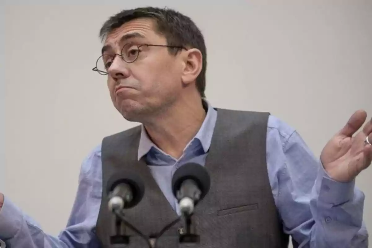 A man with glasses and a vest raises his hands in a gesture of indifference in front of two microphones.