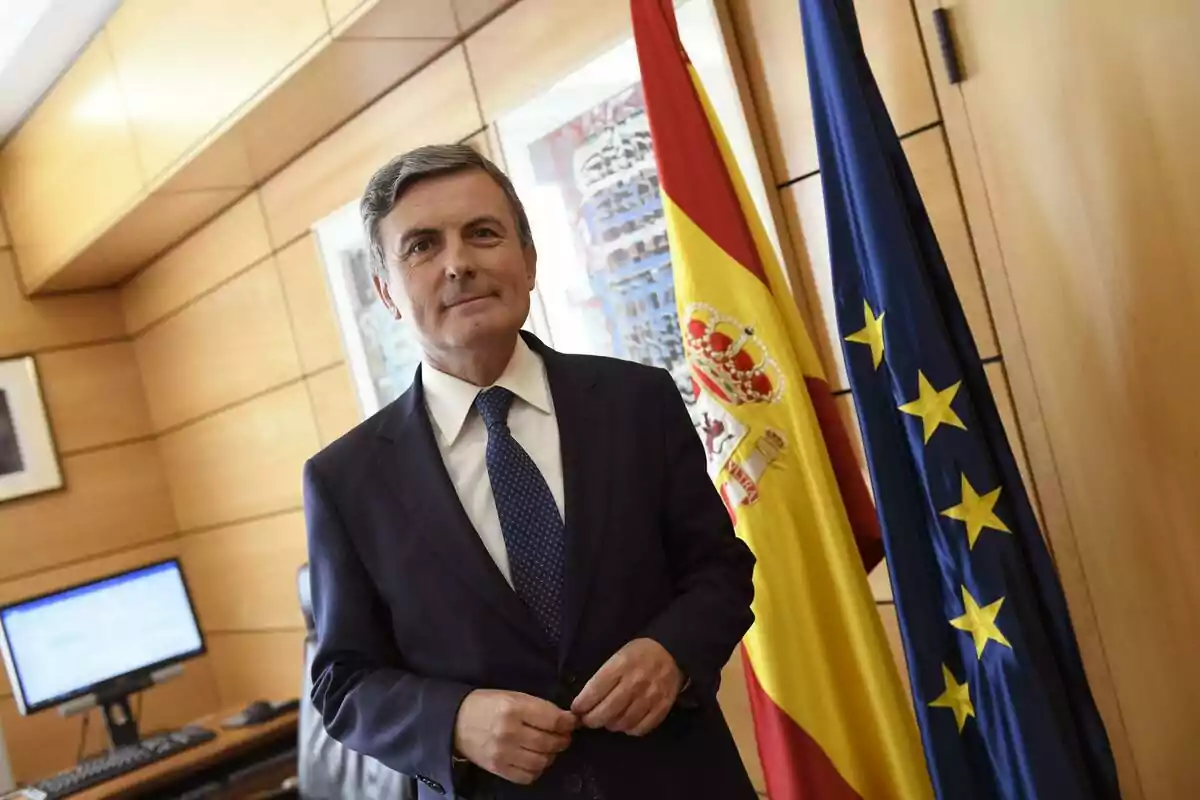 Hombre de traje posando en una oficina junto a las banderas de España y la Unión Europea