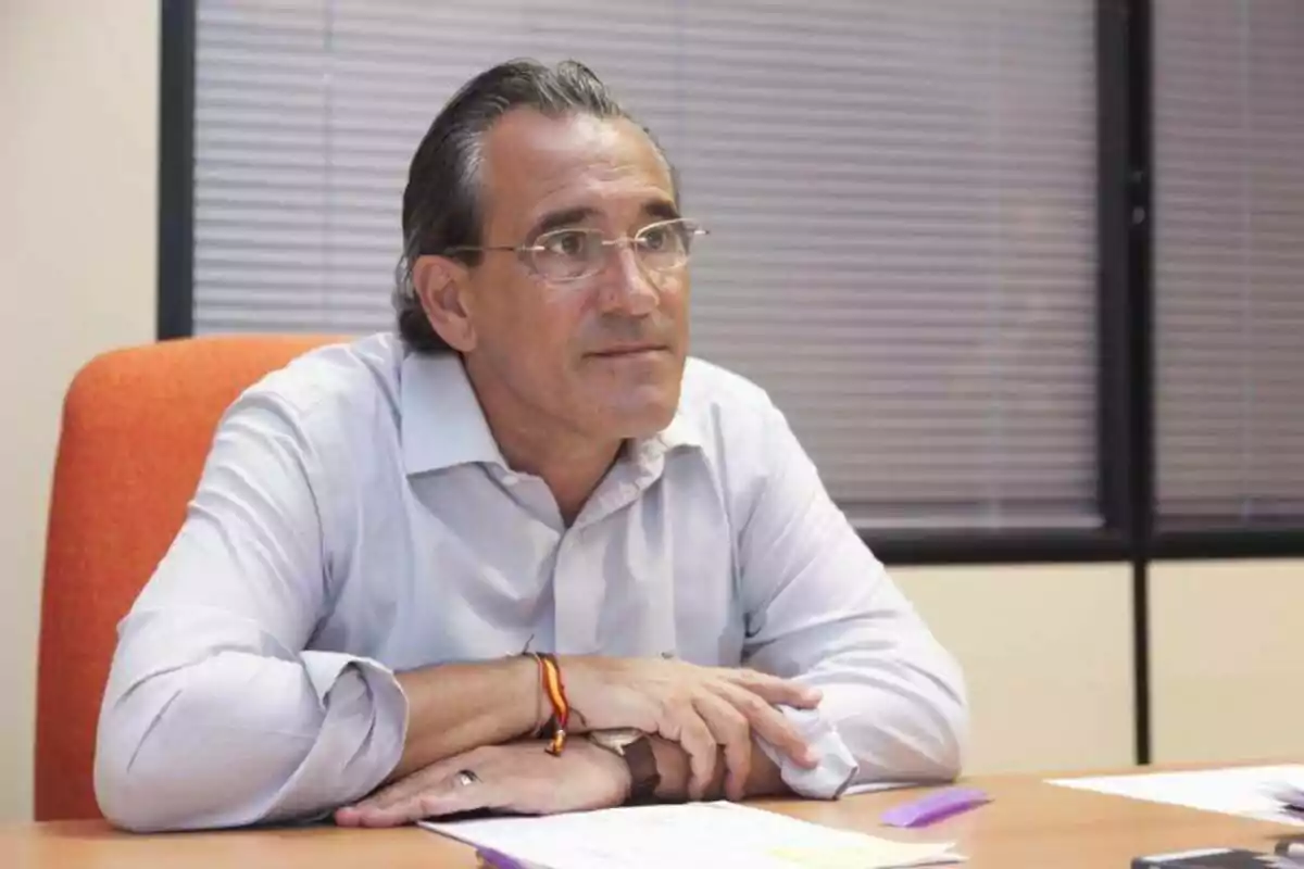 A man in a white jersey is sitting in an office with a thoughtful expression, resting his arms on a table.