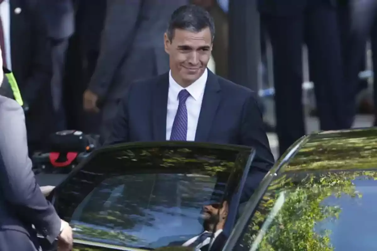 Un hombre de traje oscuro y corbata morada se encuentra junto a un automóvil con la puerta abierta, rodeado de personas en un entorno urbano.