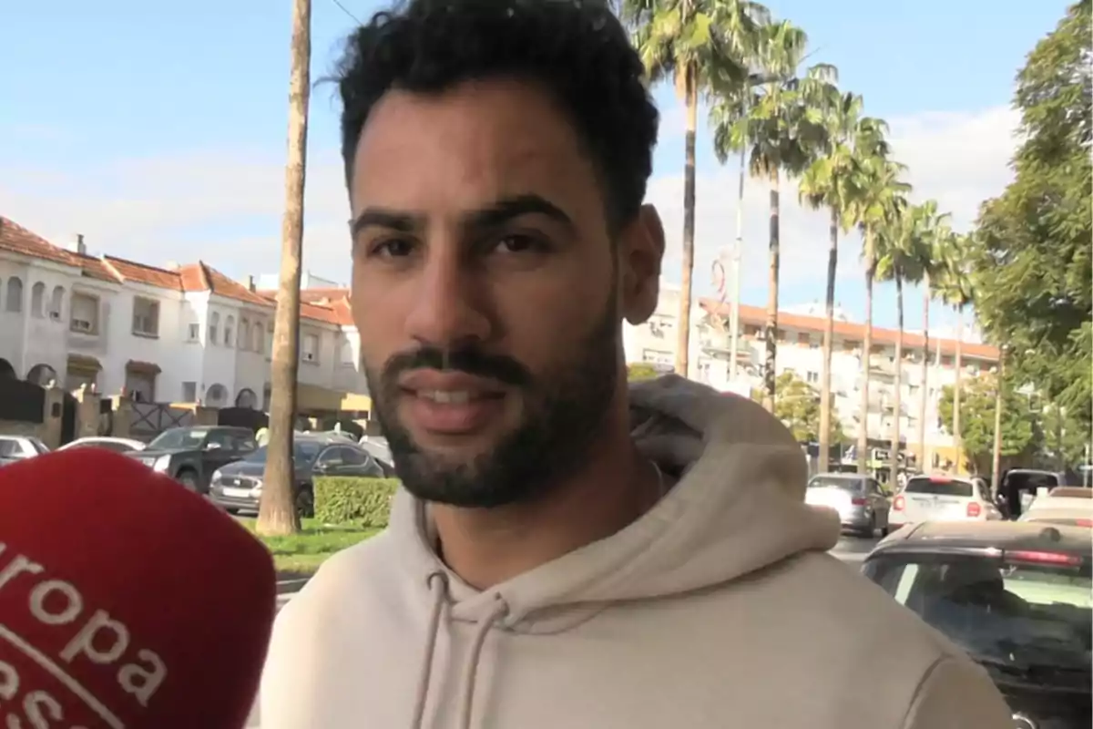 Asraf Beno con sudadera beige siendo entrevistado en la calle con palmeras y casas al fondo.