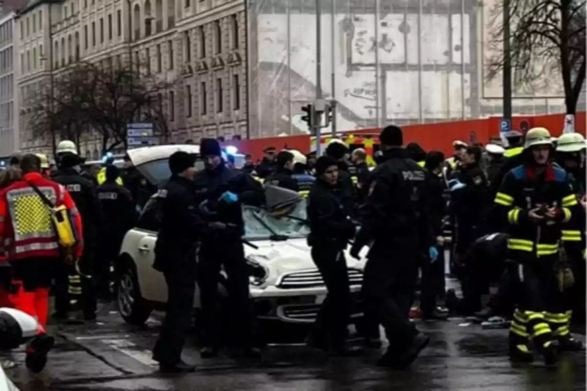 Un grupo de policías y bomberos rodea un coche blanco en una calle urbana, con edificios de fondo.