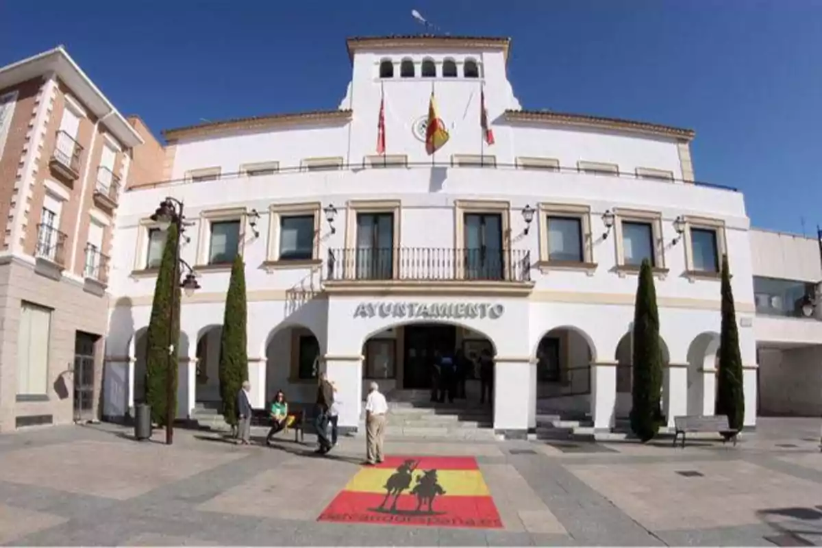 Fachada de un edificio de ayuntamiento con banderas y personas en la entrada.