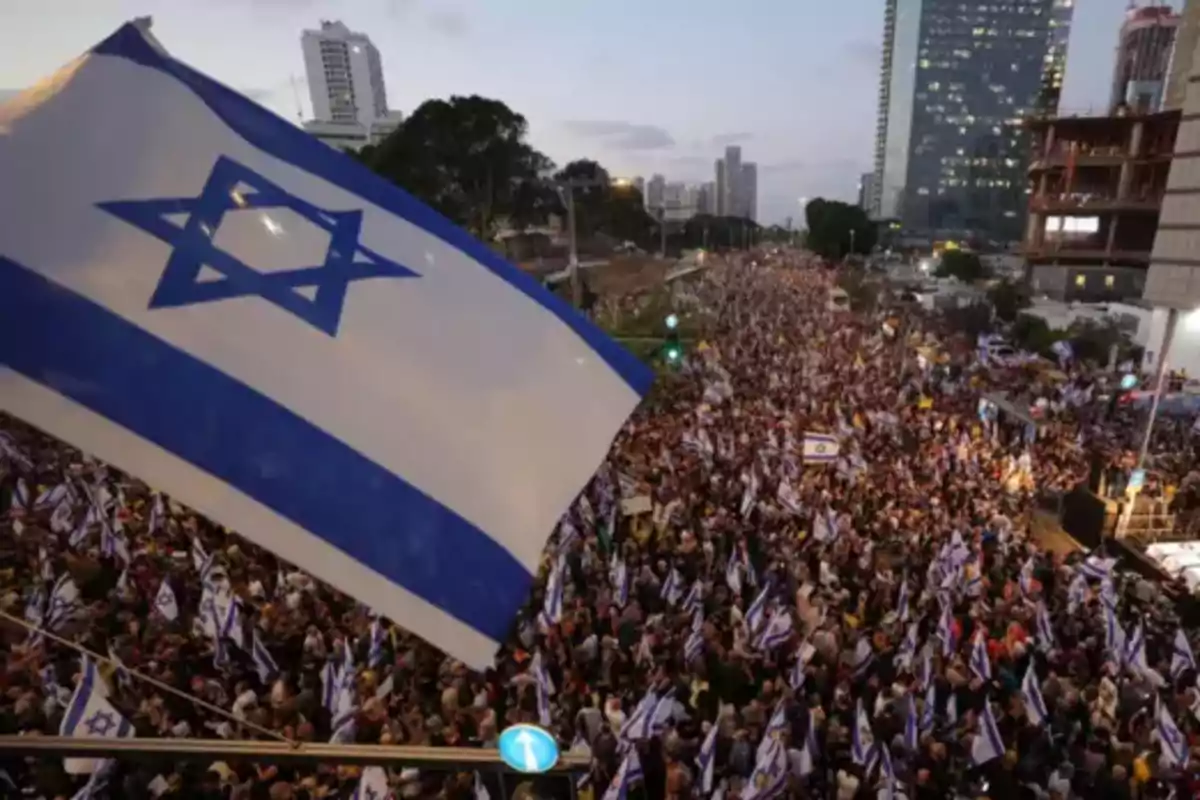 Una multitud de personas se reúne en una manifestación en una ciudad, ondeando banderas de Israel, con edificios altos al fondo.