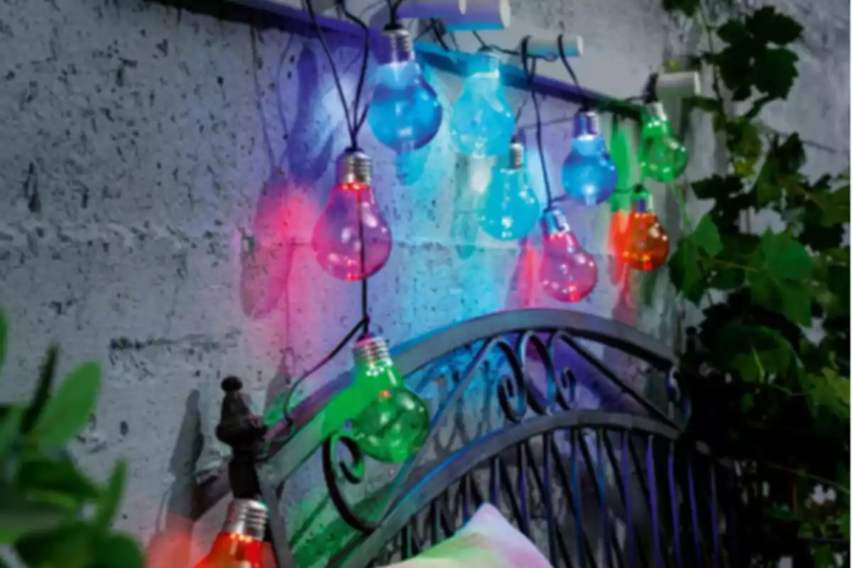 Guirnalda de focos de colores encendidos decorando una pared de cemento junto a una cama de hierro en un espacio exterior con plantas. Guirnalda de focos de colores encendidos decorando una pared de cemento junto a una cama de hierro en un espacio exterior con plantas.