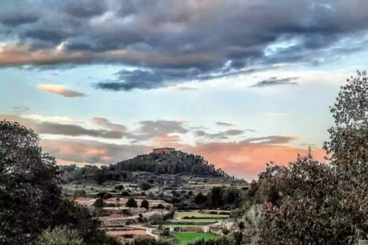 Paisaje con colina arbolada y un edificio en la cima bajo un cielo nublado con tonos rosados y azules.