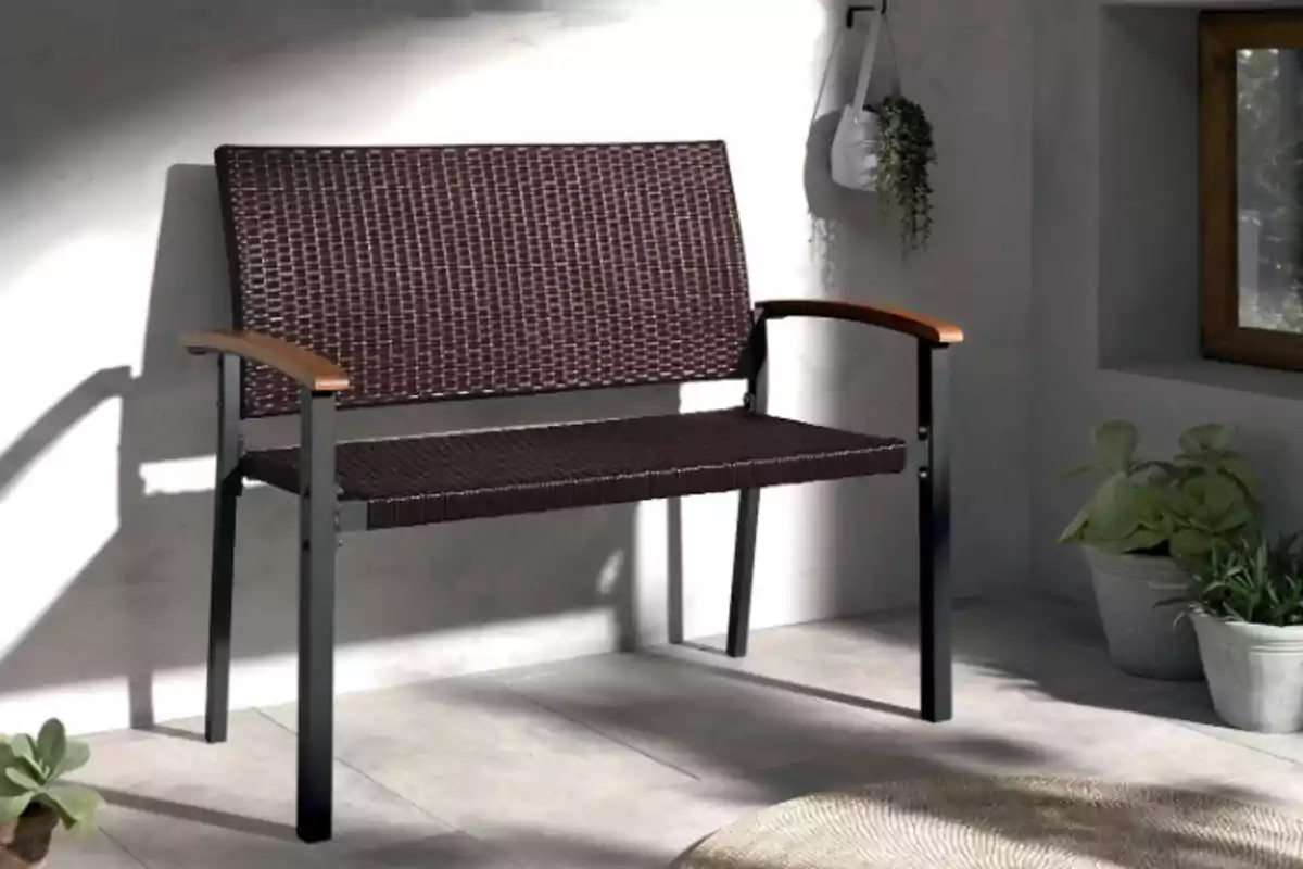 Un banco de mimbre oscuro con estructura de metal y reposabrazos de madera se encuentra en un espacio interior iluminado por la luz natural, rodeado de plantas en macetas. Un banco de mimbre oscuro con estructura de metal y reposabrazos de madera se encuentra en un espacio interior iluminado por la luz natural, rodeado de plantas en macetas.