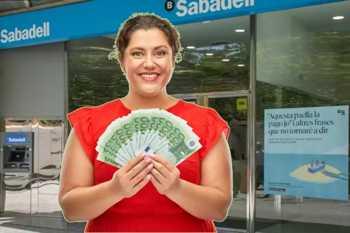 A smiling woman in a red dress holds several 100 euro notes in front of a branch of Sabadell bank.