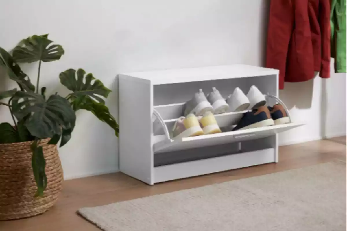 Mueble zapatero blanco con varios pares de zapatos en la entrada de una casa junto a una planta en maceta tejida y abrigos colgados en la pared Mueble zapatero blanco con varios pares de zapatos en la entrada de una casa junto a una planta en maceta tejida y abrigos colgados en la pared