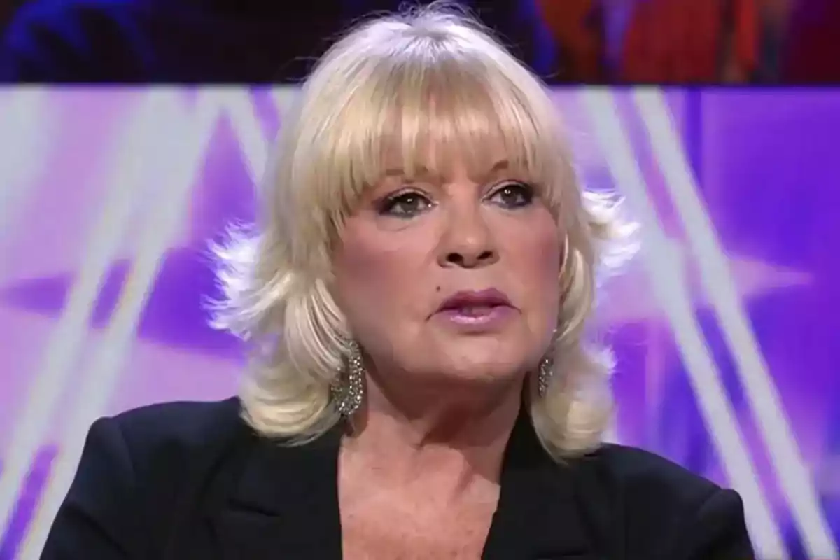 Bárbara Rey, con pendientes grandes y chaqueta negra, sentada en un set de televisión con fondo morado. Bárbara Rey, con pendientes grandes y chaqueta negra, sentada en un set de televisión con fondo morado.