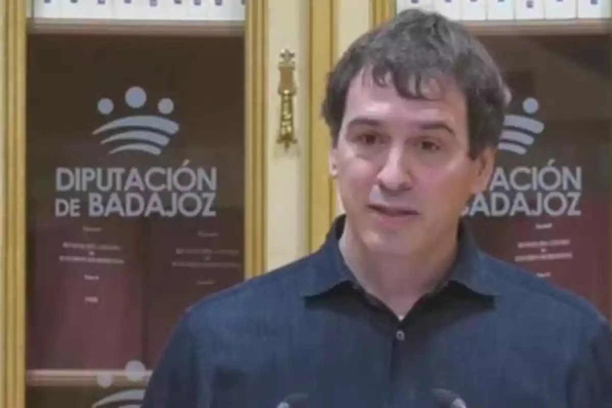 Un hombre hablando frente a un estante con libros y un cartel que dice "Diputación de Badajoz".