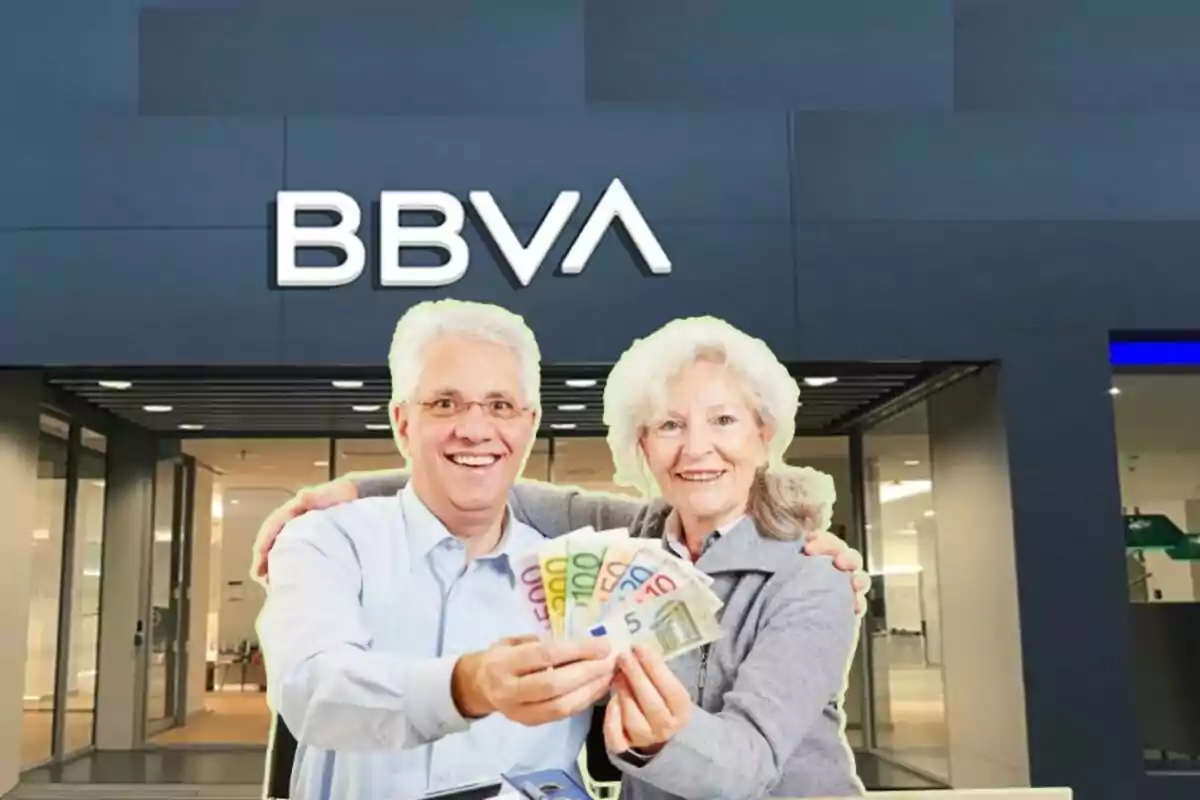 Dos personas mayores sonrientes sosteniendo billetes de euro frente a una sucursal del banco BBVA.