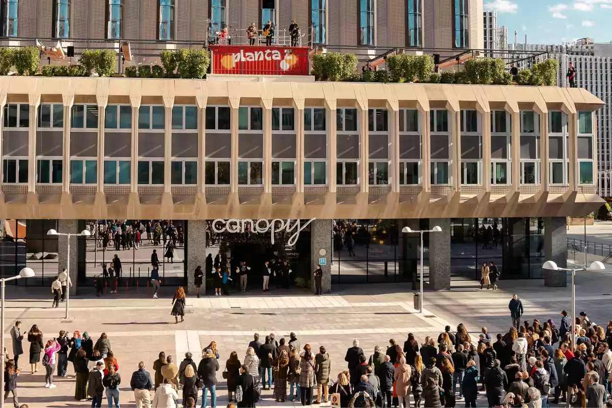 Un grupo de personas se reúne frente a un edificio moderno con un letrero que dice "canopy" mientras una banda toca en la azotea.