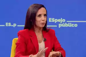 Mujer con chaqueta roja hablando en un programa de televisión con fondo azul.