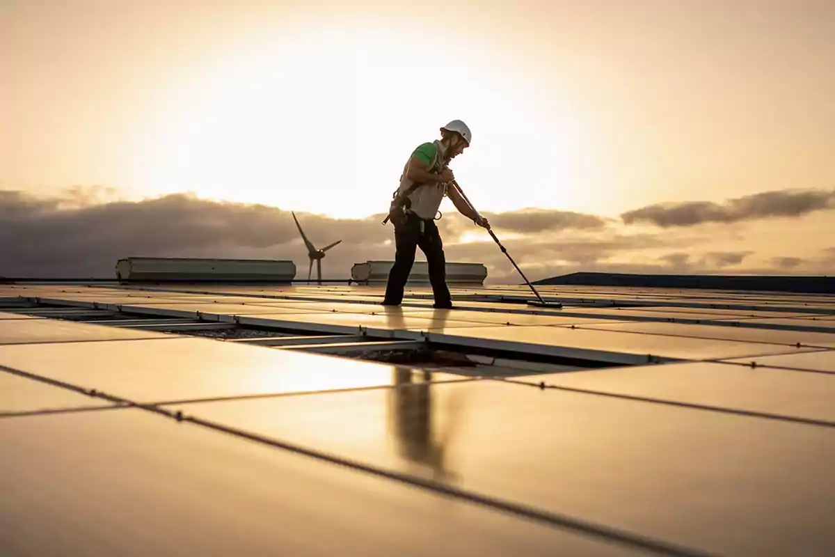 Persona limpiando paneles solares al atardecer con un aerogenerador al fondo.