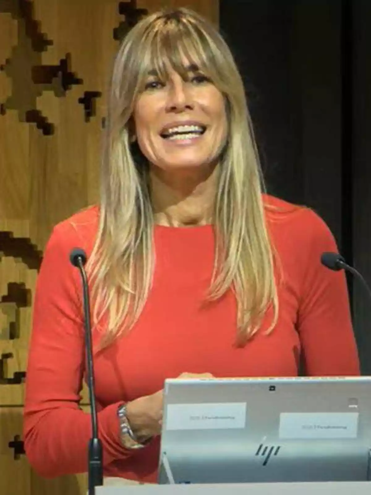 Woman with long blonde hair, wearing a red blouse, speaking in front of a microphone and a laptop.