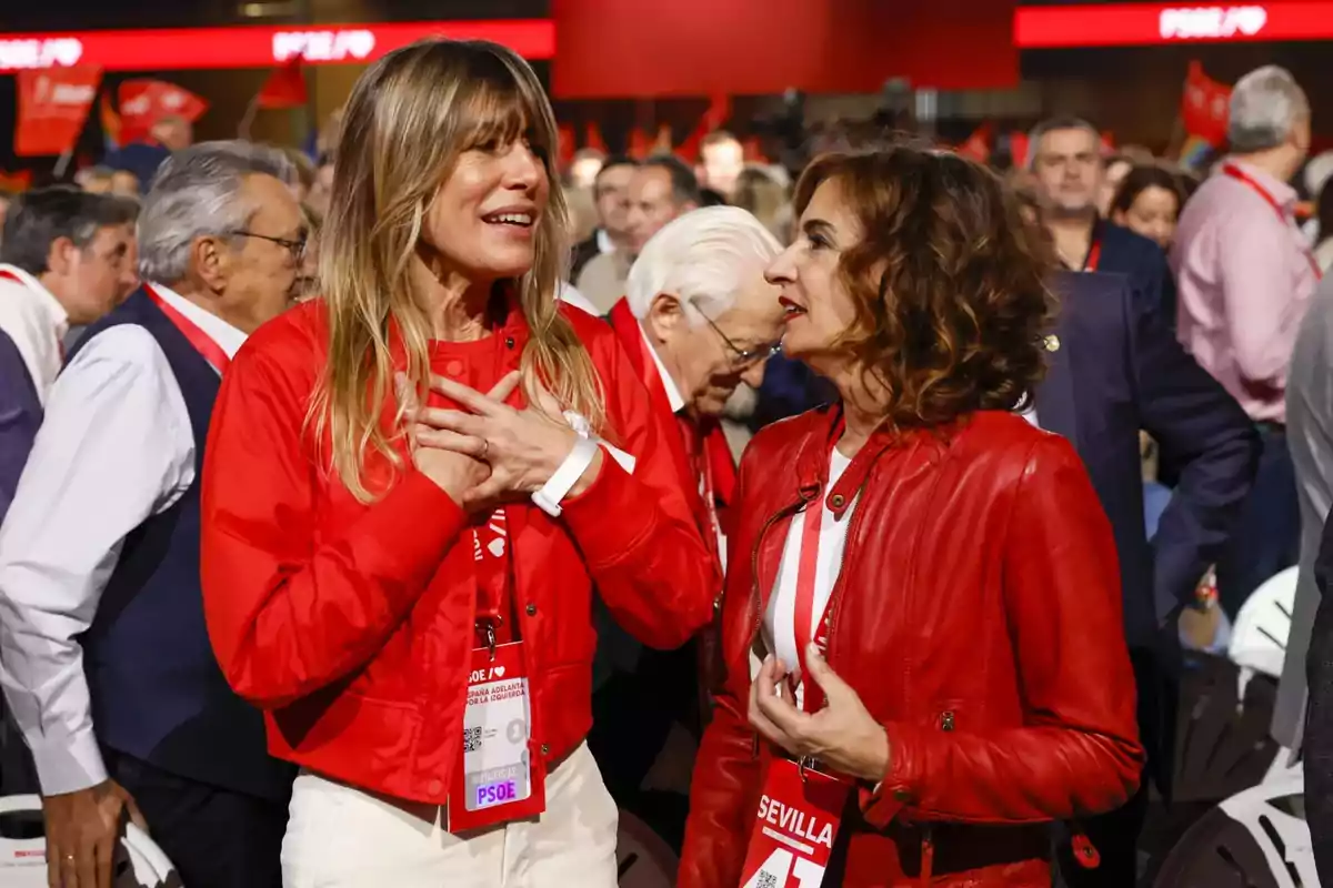 Dos mujeres conversan animadamente en un evento, rodeadas de personas, ambas visten chaquetas rojas y llevan acreditaciones colgadas al cuello. Dos mujeres conversan animadamente en un evento, rodeadas de personas, ambas visten chaquetas rojas y llevan acreditaciones colgadas al cuello.