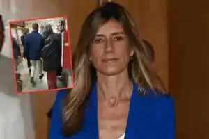 Una mujer con cabello largo y rubio lleva un blazer azul mientras camina por un pasillo; en la esquina superior izquierda hay una imagen insertada de varias personas caminando por un pasillo.