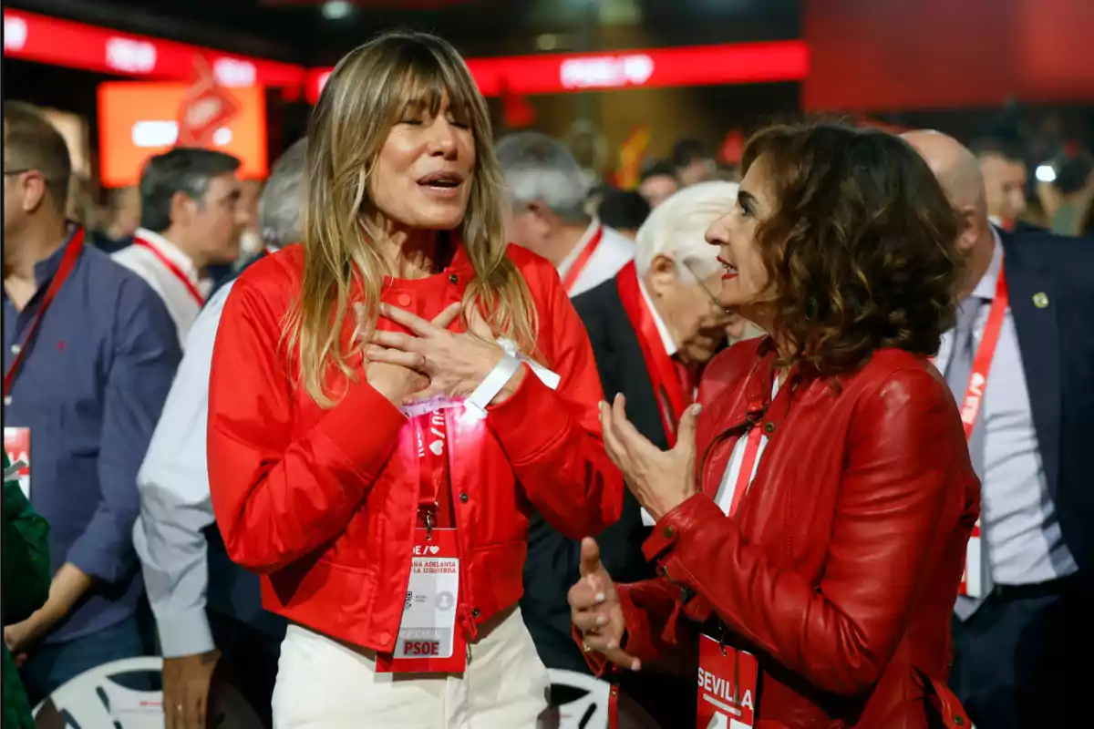 Dos mujeres conversan animadamente en un evento político, ambas visten chaquetas rojas y llevan acreditaciones colgadas al cuello, rodeadas de otras personas en un ambiente concurrido. Dos mujeres conversan animadamente en un evento político, ambas visten chaquetas rojas y llevan acreditaciones colgadas al cuello, rodeadas de otras personas en un ambiente concurrido.