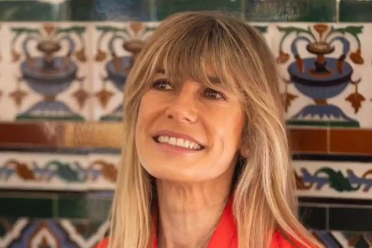 Una mujer sonriente con cabello rubio y flequillo, vestida con una camisa roja, está frente a un fondo de azulejos decorativos con patrones coloridos.