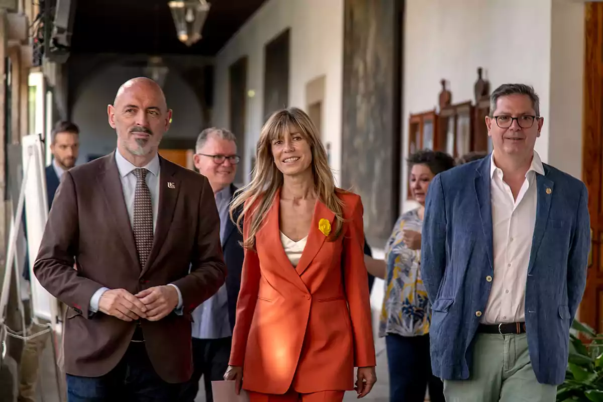 Un grupo de personas caminando por un pasillo, con tres personas en primer plano, una mujer en el centro con un traje naranja y dos hombres a los lados, uno con un traje marrón y el otro con un traje azul.