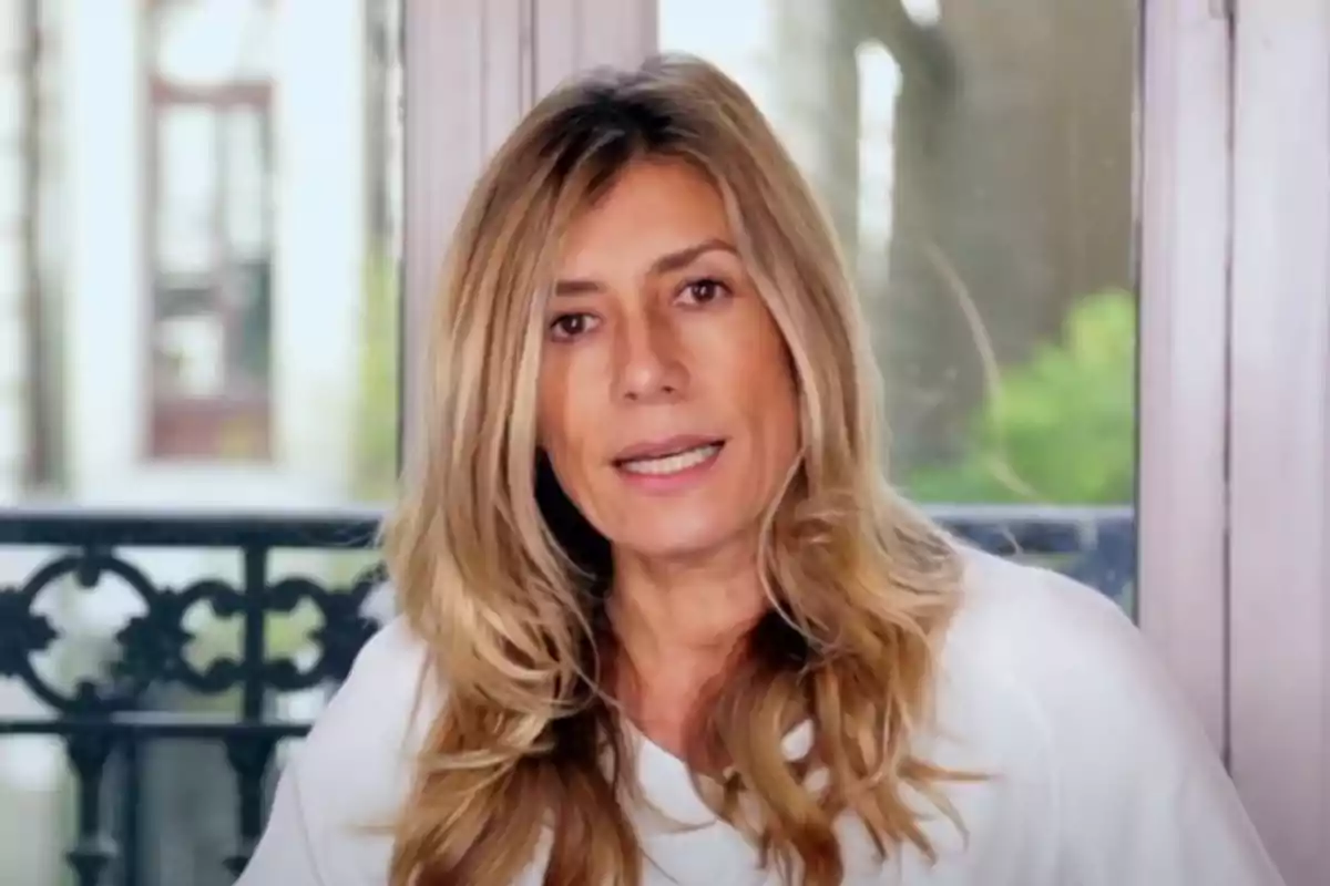 Una mujer de cabello rubio y largo, vestida con una blusa blanca, está sentada frente a una ventana con un fondo borroso.