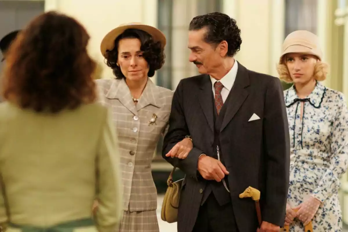 A group of people dressed in period clothing in an interior scene, where a woman appears excited while a man walks by her arm and another woman watches from behind.