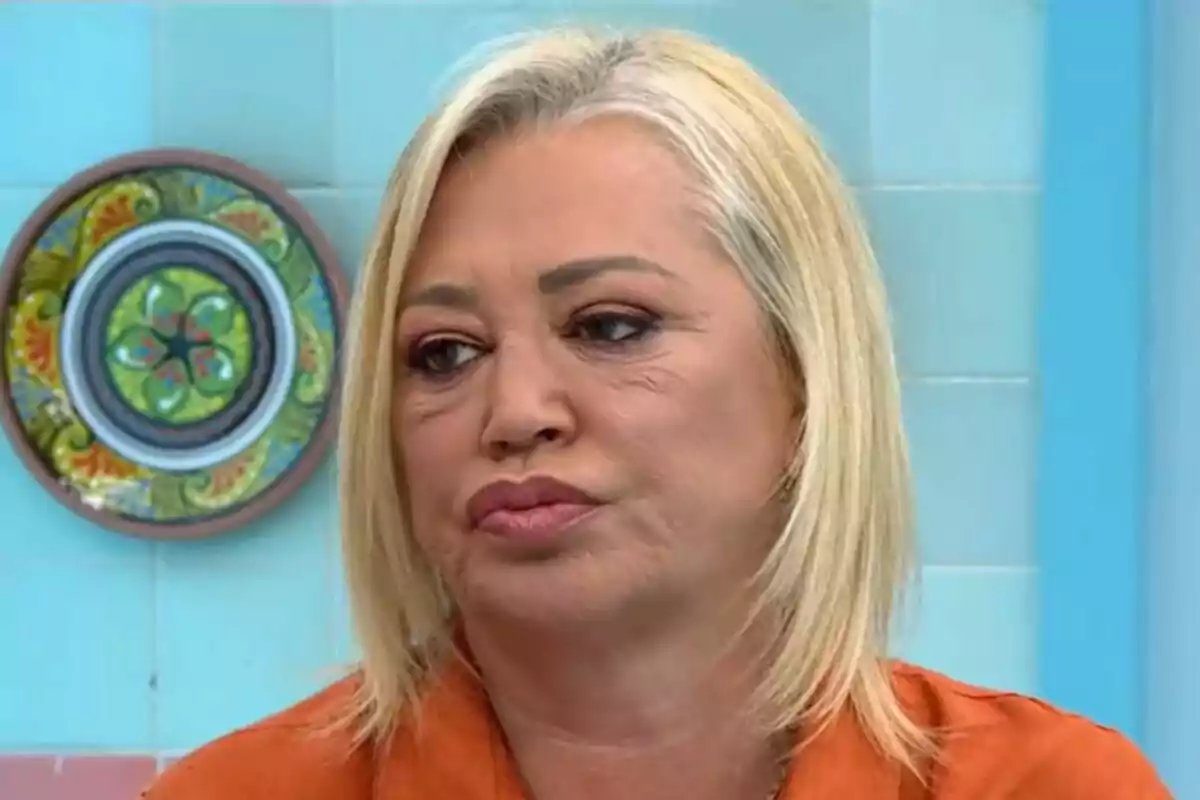 Belén Esteban de cabello rubio y expresión seria en un entorno con azulejos azules y un plato decorativo en la pared en La Familia de la Tele. Belén Esteban de cabello rubio y expresión seria en un entorno con azulejos azules y un plato decorativo en la pared en La Familia de la Tele.