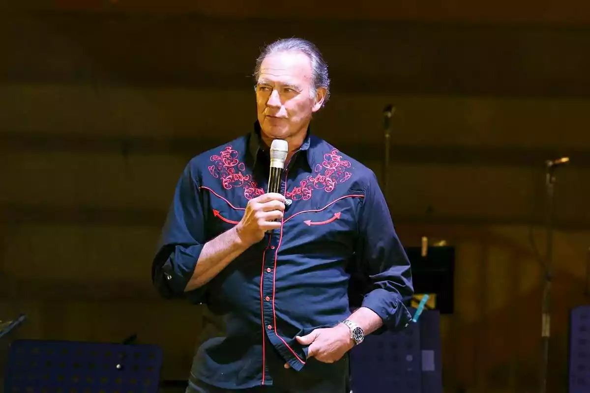 Bertín Osborne hablando mirando a un lado con camisa azul y roja y un micrófono en la mano