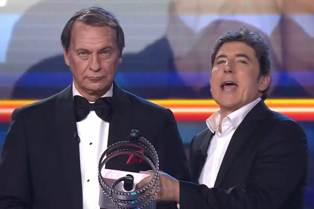 Dos hombres en un escenario, Manel Fuentes sostiene un objeto circular mientras Bertín Osborne lleva un esmoquin y corbata de moño en Tu Cara Me Suena. Dos hombres en un escenario, Manel Fuentes sostiene un objeto circular mientras Bertín Osborne lleva un esmoquin y corbata de moño en Tu Cara Me Suena.