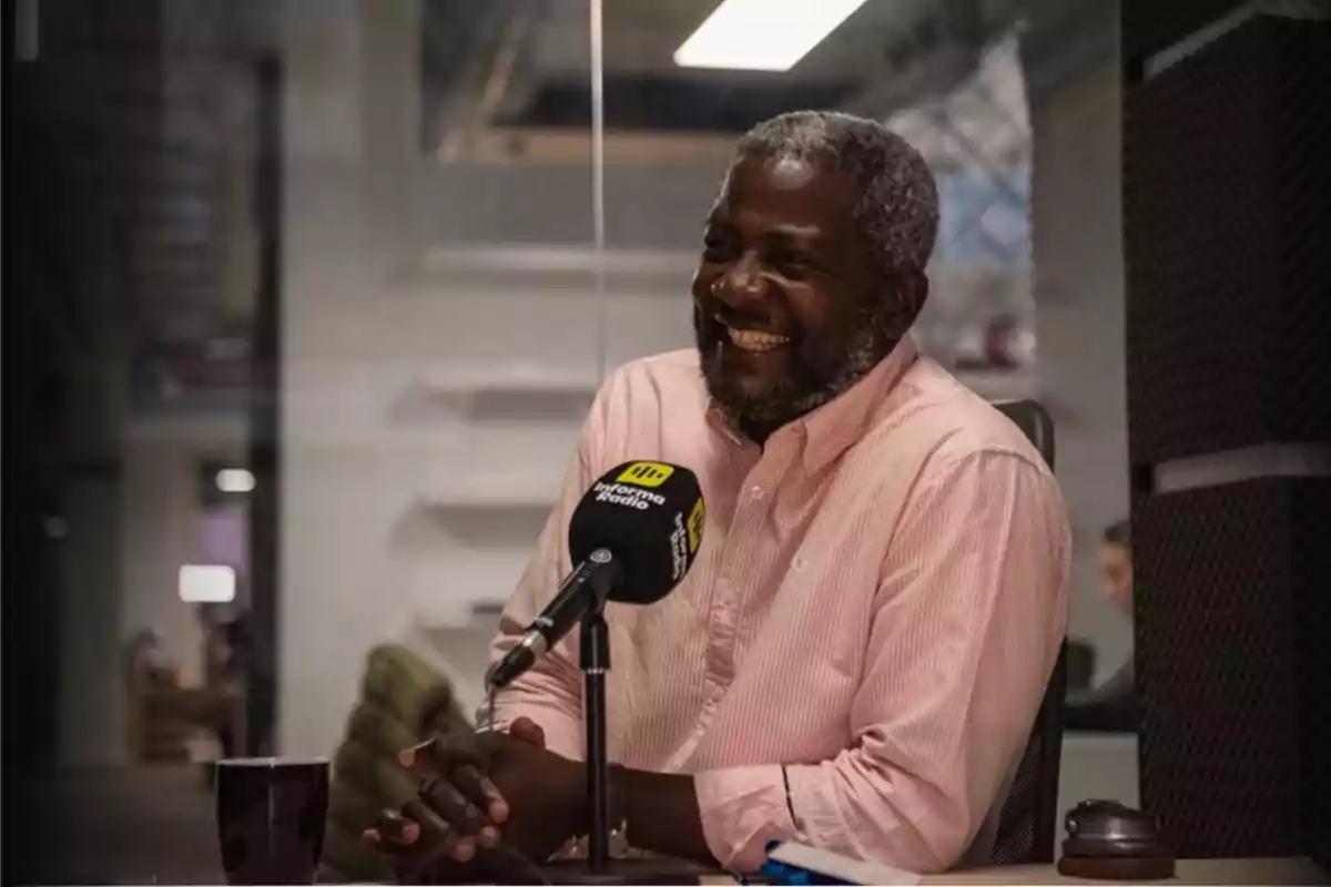 Un hombre sonriente con camisa rosa está sentado frente a un micrófono en un estudio de radio.