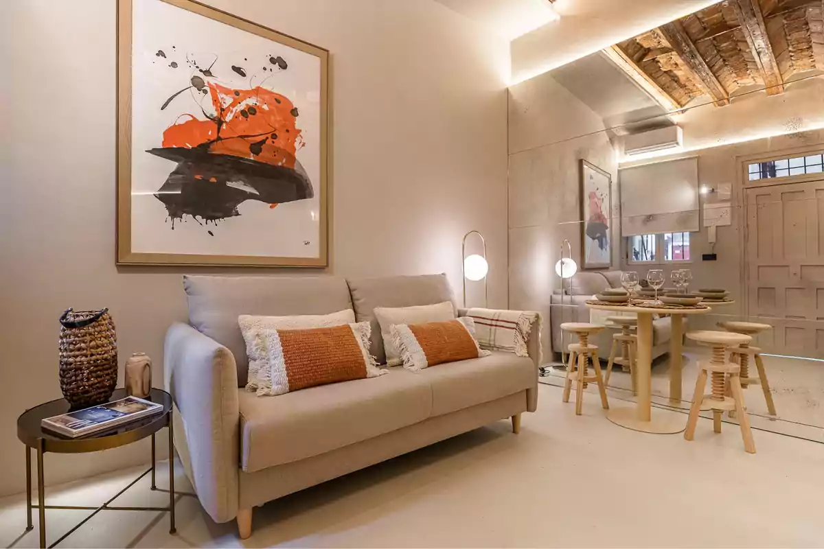 Una sala de estar moderna con un sofá beige decorado con cojines naranjas y blancos, una mesa lateral con revistas y un jarrón tejido, una pintura abstracta en la pared, una mesa de comedor con platos y copas, y un techo de vigas de madera expuestas.