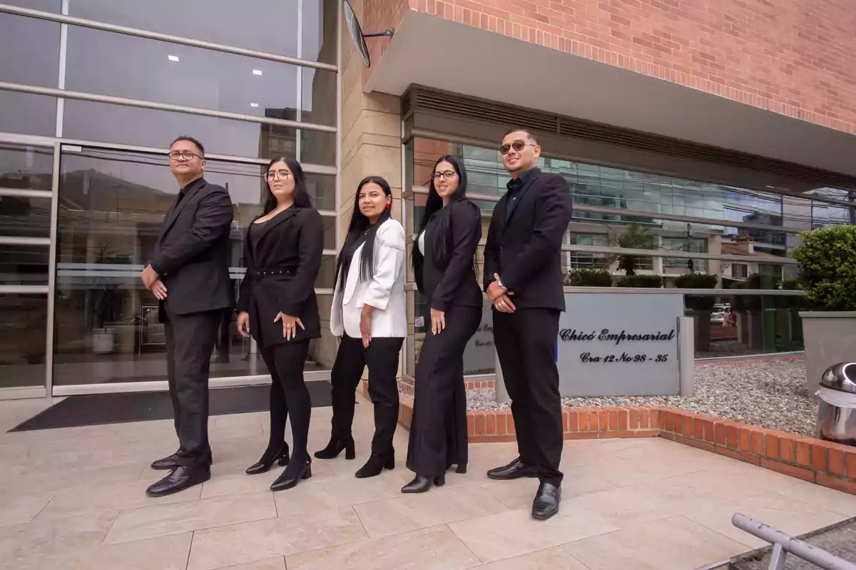 Cinco personas vestidas de manera formal posan frente a un edificio de oficinas con fachada de vidrio y ladrillo
