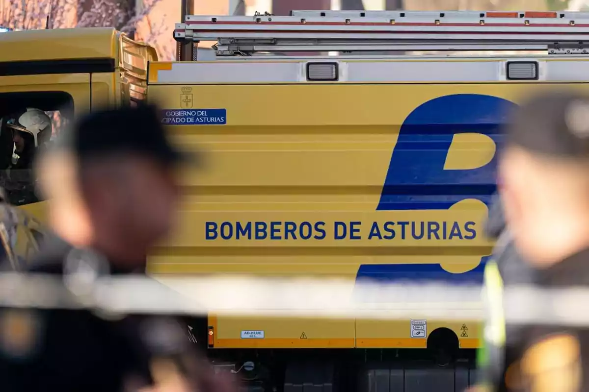 Camión amarillo de los bomberos de Asturias con personal de emergencia y policías en primer plano desenfocados