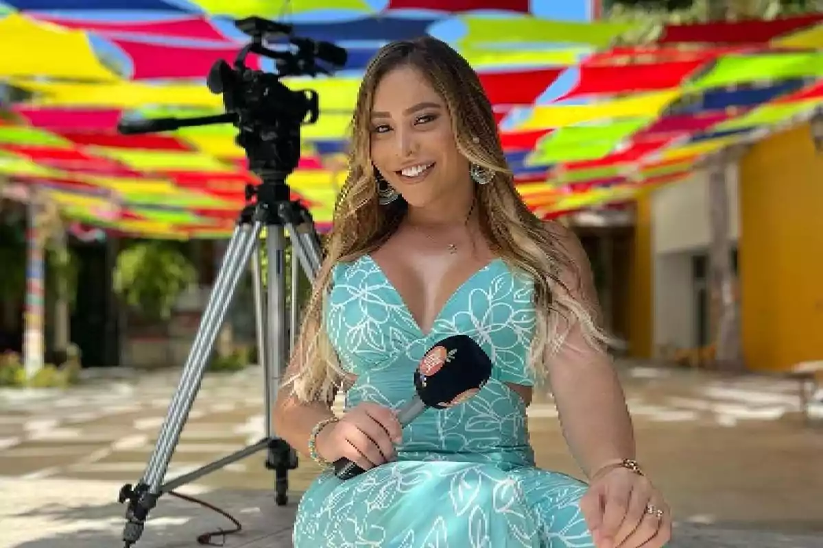 Mujer con vestido azul sosteniendo un micrófono, sentada frente a una cámara de video bajo un techo de colores brillantes.