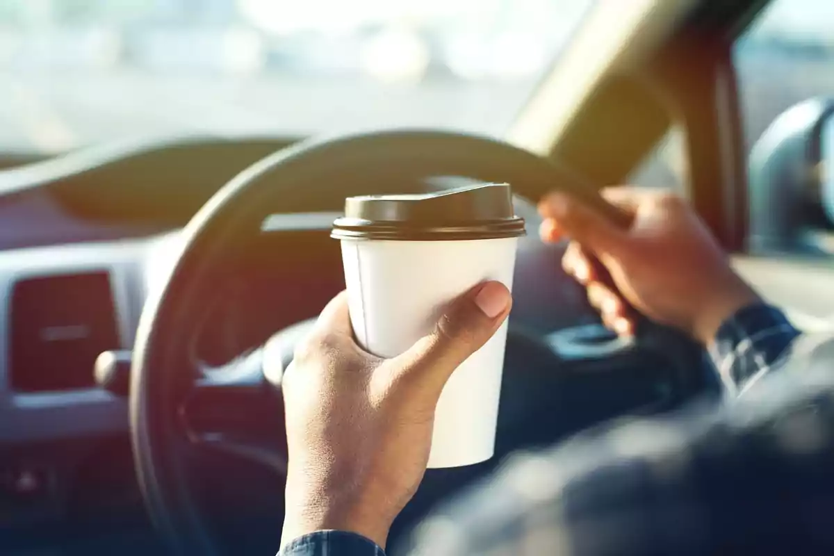 Persona conduciendo un automóvil mientras sostiene un vaso de café desechable.