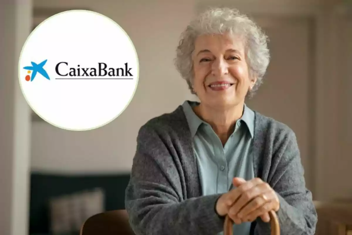 Una mujer mayor sonriente con cabello canoso y un suéter gris, apoyada en un bastón, con el logo de CaixaBank en la esquina superior izquierda. Una mujer mayor sonriente con cabello canoso y un suéter gris, apoyada en un bastón, con el logo de CaixaBank en la esquina superior izquierda.