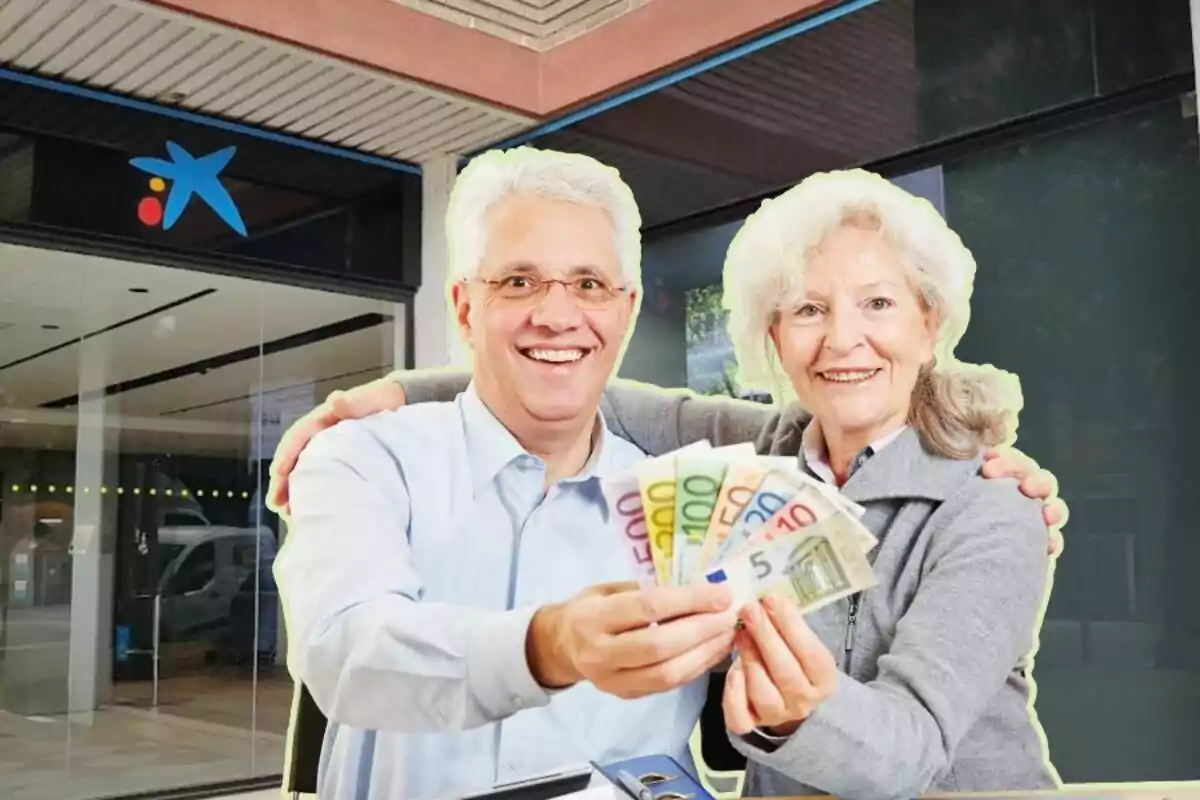 Dos personas mayores sonrientes sosteniendo billetes de euro frente a una sucursal bancaria.