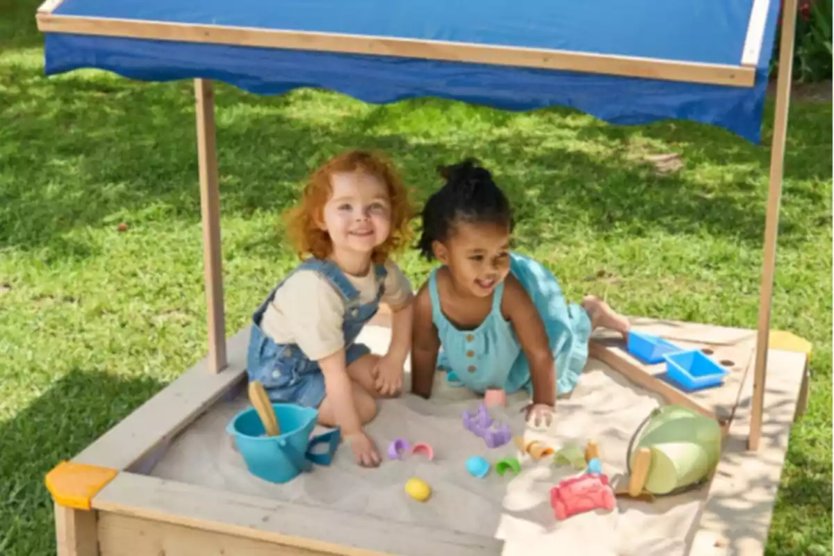 Dos niñas pequeñas juegan felices en una caja de arena bajo una sombrilla azul rodeadas de juguetes de playa en un jardín verde Dos niñas pequeñas juegan felices en una caja de arena bajo una sombrilla azul rodeadas de juguetes de playa en un jardín verde