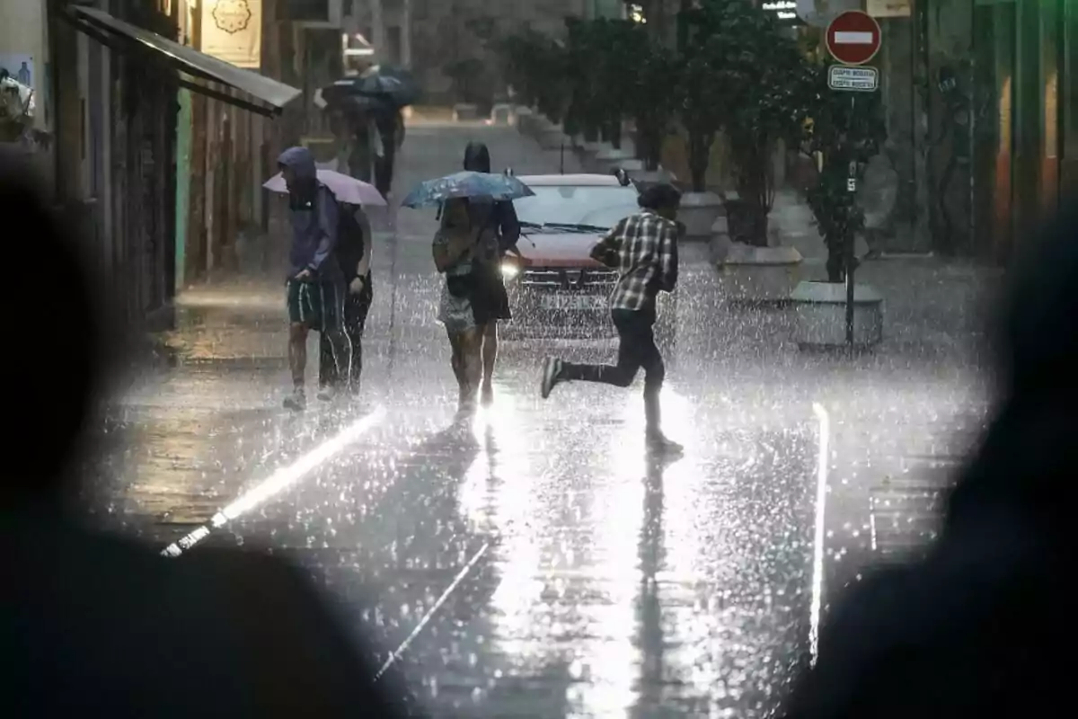 Imagen de una calle mientras llueve con personas corriendo, otras con paraguas y un coche circulando