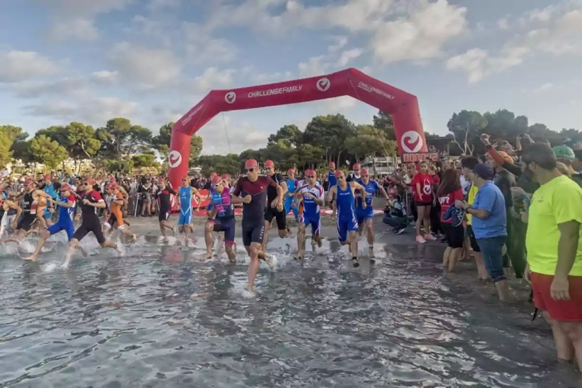 Un grupo de atletas comienza una carrera de triatlón desde la playa, rodeados de espectadores y bajo un arco inflable rojo.