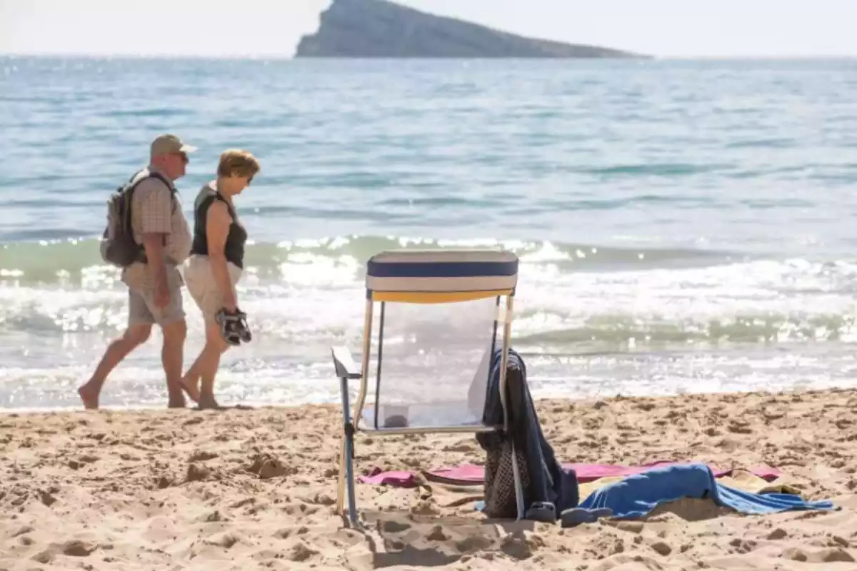 Dos personas mayores pasean a la orilla de la playa, con una silla vacía en la arena y el mar de fondo