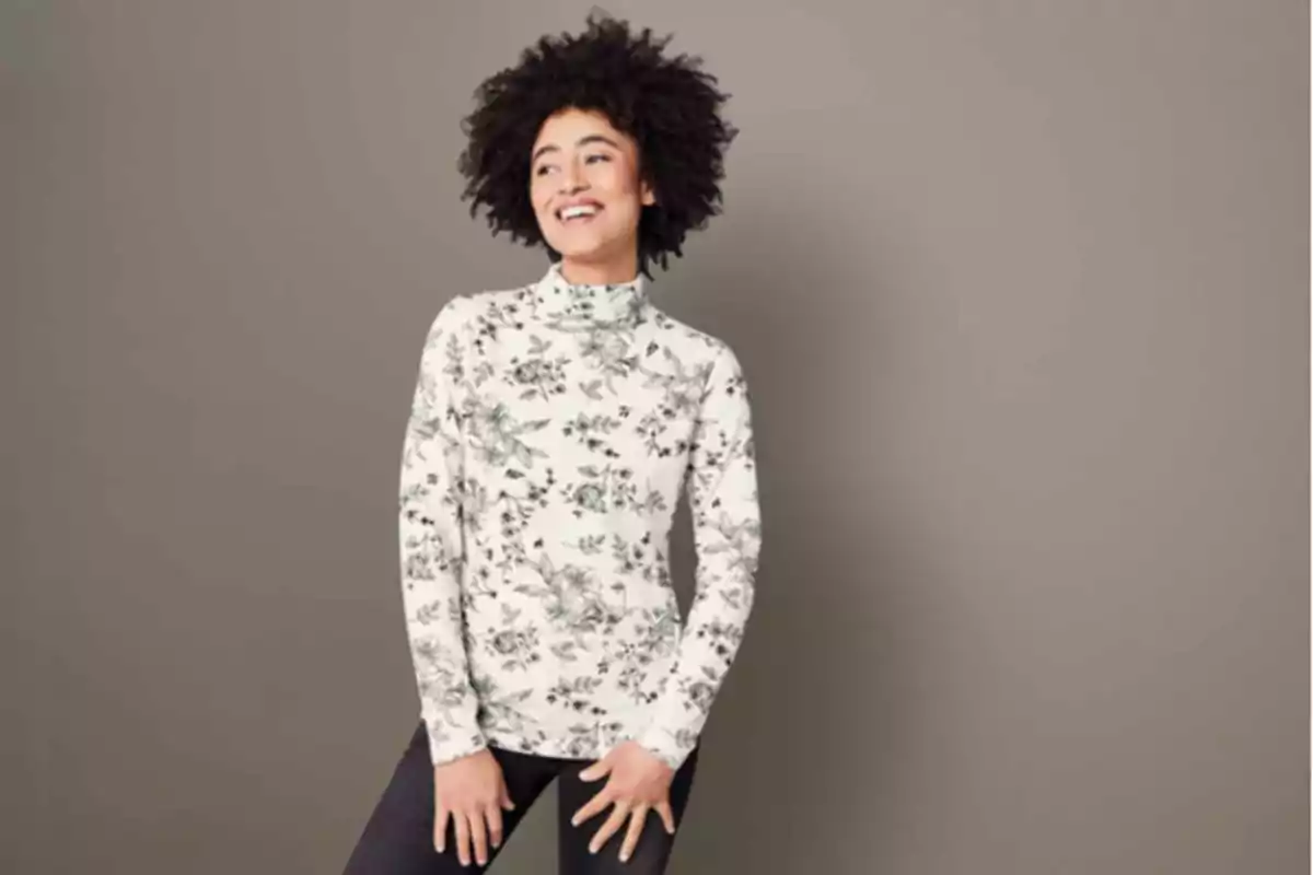 Mujer sonriente con cabello rizado usando un suéter de cuello alto con estampado floral en un fondo gris. Mujer sonriente con cabello rizado usando un suéter de cuello alto con estampado floral en un fondo gris.