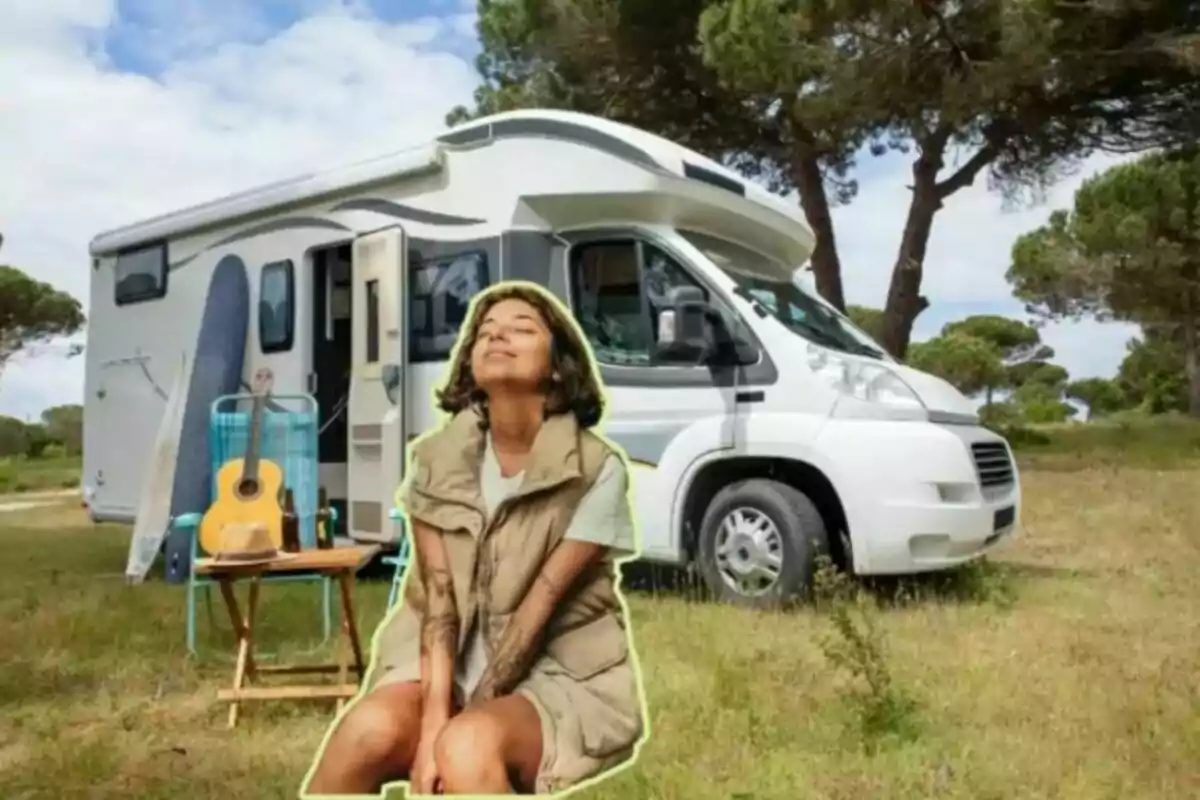 Una mujer sentada en el césped frente a una autocaravana, con una guitarra y una mesa pequeña al lado.