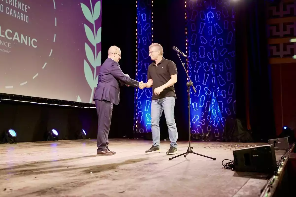 Dos personas se dan la mano en un escenario iluminado con un micrófono al frente y un fondo decorativo.