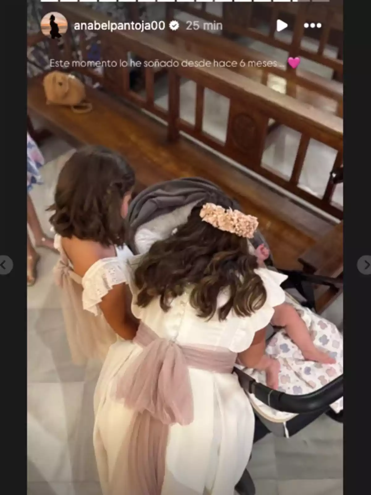 Dos niñas con vestidos blancos y lazos rosados miran a un bebé en un cochecito dentro de una iglesia Dos niñas con vestidos blancos y lazos rosados miran a un bebé en un cochecito dentro de una iglesia