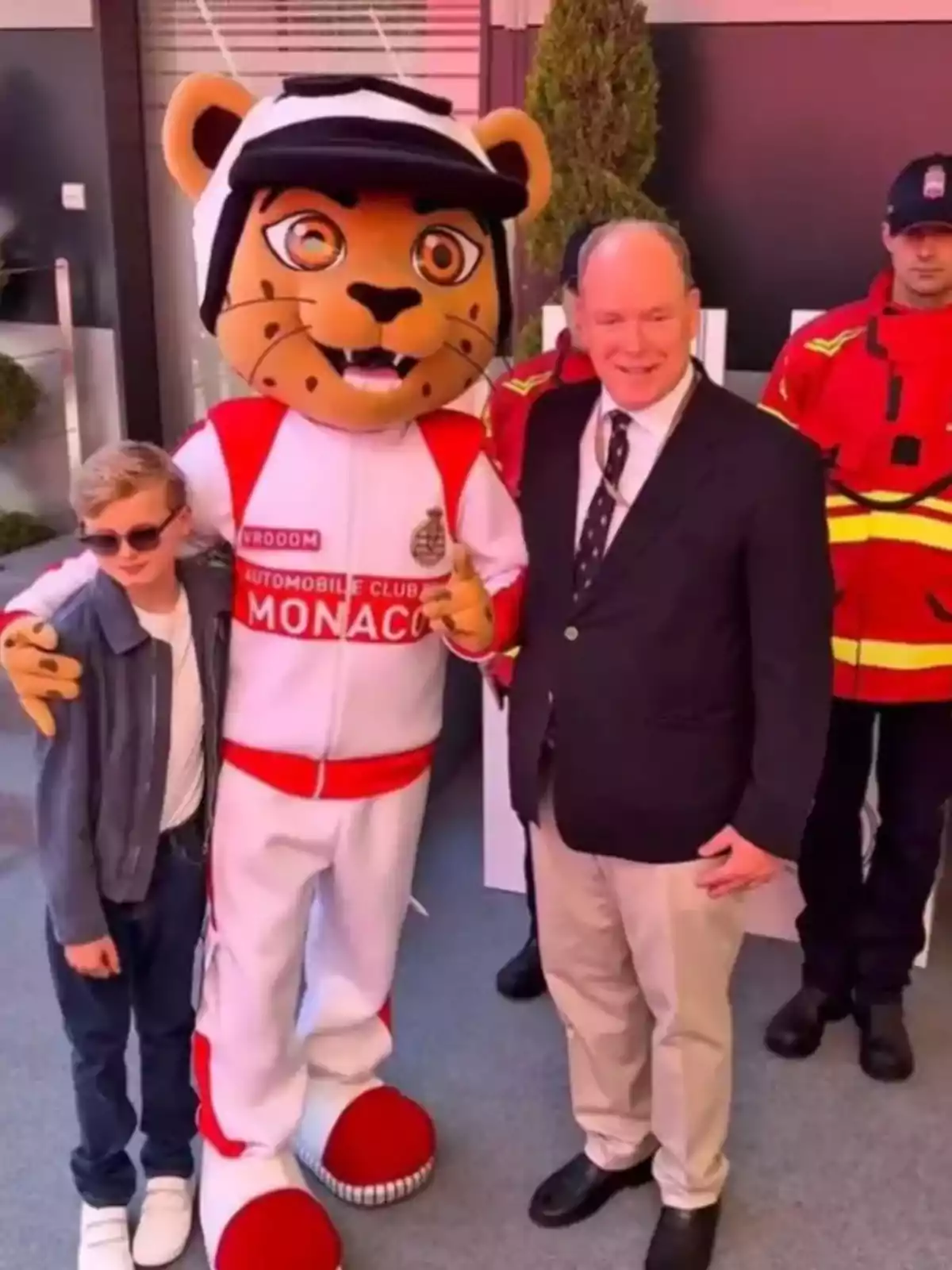 Jacques with sunglasses, a person dressed as a tiger in an Automobile Club of Monaco uniform, and Alberto of Monaco in a suit pose together while two people in red uniforms stand behind them. Jacques with sunglasses, a person dressed as a tiger in an Automobile Club of Monaco uniform, and Alberto of Monaco in a suit pose together while two people in red uniforms stand behind them.