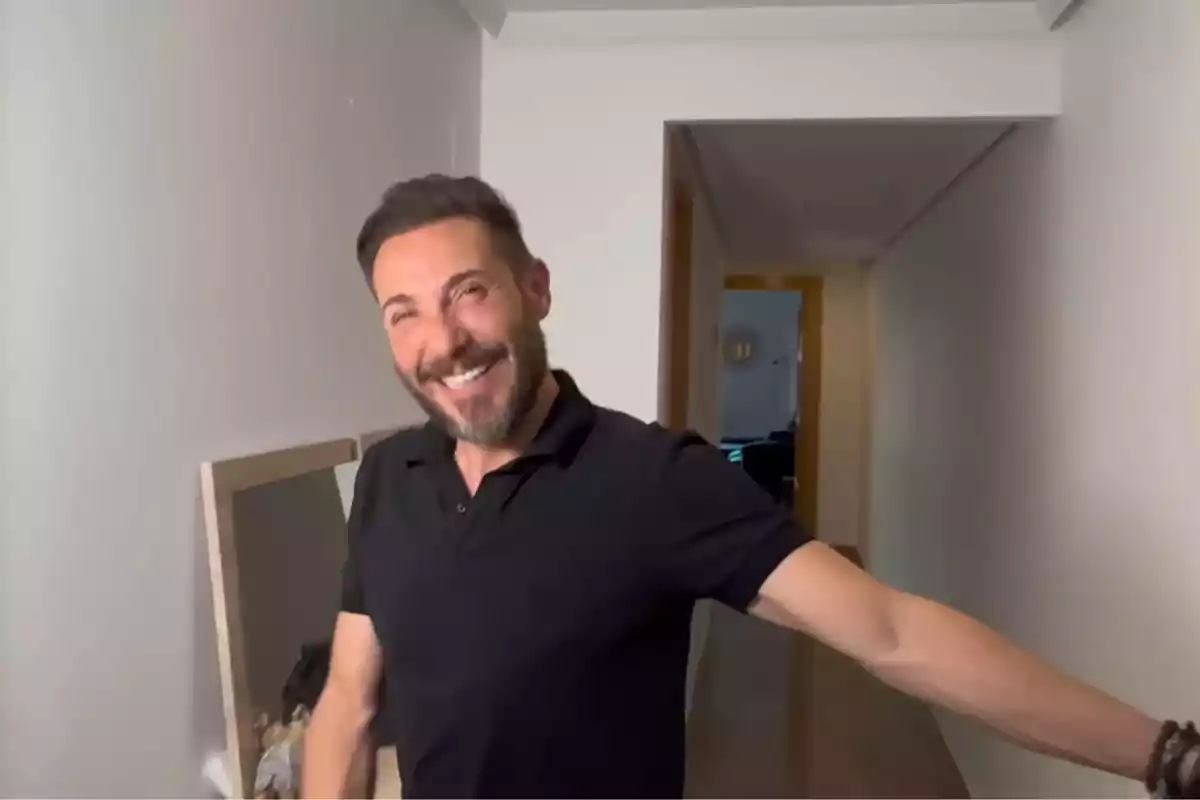 Antonio David Flores sonriente con barba y camiseta negra posando en un pasillo de una casa Antonio David Flores sonriente con barba y camiseta negra posando en un pasillo de una casa